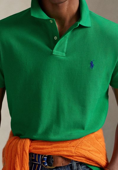 Πράσινο πόλο πουκάμισο με υφαντό υφή, με μπλε λογότυπο, συνοδευόμενο από ένα πορτοκαλί πλεκτό πουλόβερ δεμένο γύρω από τη μέση.