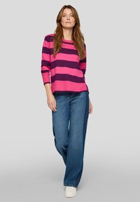 Pullover mit pinken und tiefvioletten Streifen und dreiviertellangen Ärmeln, kombiniert mit hellblauen Jeans mit weitem Bein und weißen Sneakern. Glatte Textur.