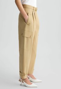 Pantalons cargo beiges avec poches latérales et accents de boutons, dotés d'une taille ceinturée et de revers, associés à des escarpins blancs à bout pointu.