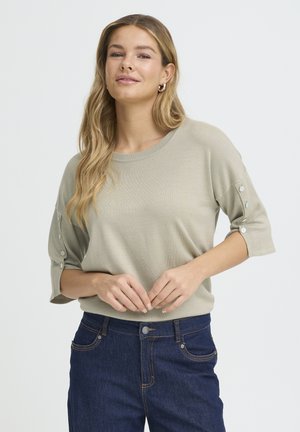 Jasnozielony sweter z okrągłym dekoltem, krótkimi rękawami i ozdobnymi guzikami na rękawach. Zestawiony z ciemnoniebieskimi dżinsami. Gładka tekstura.