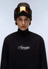 Fekete, hosszú ujjú póló, álló nyakkal, amelyen fehér, kézírásos "Napapijri" logó található. A megjelenést egy barna kötött sapka és tükröződő szemüveg egészíti ki.