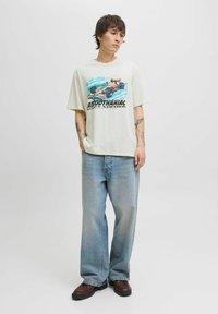 Tricou bej deschis cu un grafic imprimat al unei mașini în nuanțe de albastru și portocaliu. Asortat cu blugi evazați de un albastru estompat și pantofi sport maro.