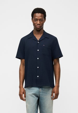 Mlajši moški, oblečen v temnomodro kratkoročno srajco z gumbi in prsnim žepom ter svetlo modre kavbojke, stoji pred enobarvnim ozadjem.