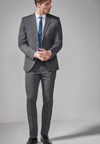 Costume gris composé d'un blazer ajusté et de pantalons droits, associé à une chemise blanche et une cravate bleue. Tissu lisse avec un éclat subtil.