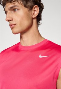 Różowa bezrękawnikowa koszulka sportowa z okrągłym dekoltem, wykonana z gładkiego materiału. Posiada niewielkie białe logo Nike na górnej części lewej piersi.