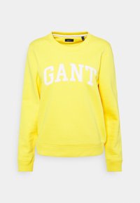 Sudadera amarilla de cuello redondo, fabricada con un tejido suave, que presenta un gran logo blanco "GANT" en el pecho y puños y bajo acanalados.