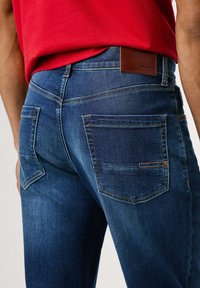 Temno modri kavbojki z oblikovanim krojem, dvema zadnjima žepoma in rjavim usnjenim logotipom na pasu. Gladka tekstura.