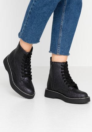 Sort glitrende ankelstøvler med snørebånd og sideskuldre, iført sammen med cropped blå denim jeans på hvidt gulv.