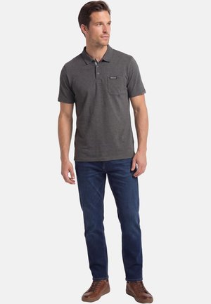 Mann trägt dunkelgraues, kurzärmeliges Poloshirt mit Brusttasche, blaue Jeans und braune Ledersneaker, steht und schaut nach rechts.