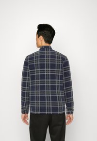 Camicia flanella a motivi a quadri blu navy con linee grigie e olive, colletto classico, maniche lunghe e chiusura a bottoni sui polsini. Dettaglio di cucitura visibile.