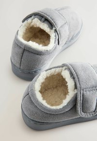 Chaussons gris avec un intérieur doux et moelleux et une sangle en Velcro. L'extérieur est en tissu texturé, avec un bout arrondi et une semelle robuste.