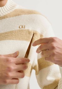Gestreifter Strickpullover in Creme und Beige mit einem gerippten Kragen, der eine sichtbare Öffnung an der Schulter und ein kleines Logo aufweist.