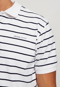 Biała koszulka polo z niebieskimi poziomymi paskami, krótkie rękawy i kołnierzyk. Zawiera logo "CROSS JEANS" na lewej piersi.
