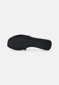 Semelle de chaussure noire à bout carré, talon bas, grip texturé et quatre patins antidérapants ronds, présentée sur un fond blanc uni.
