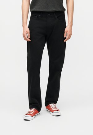 Mand iført sorte jeans med lige ben og røde lave sneakers med hvide snørebånd, stående mod en ensfarvet lys baggrund.