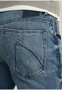 Jeans de denim azul que presentan un diseño distintivo de bolsillos traseros en ángulo y un parche de cuero con la marca en la cinturilla. Textura suave.