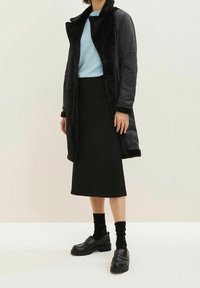 Mustapituinen pitkä takki tekoturkishaalarilla, vaaleansininen paita, musta kynähame ja paksupohjaiset mustat loafer-kengät.
