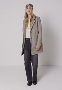 Femme portant un manteau gris, un haut blanc, un jean noir, des chaussures noires et un bandana imprimé léopard, debout devant un fond clair uni.
