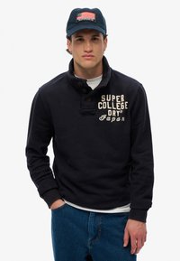 Maglione blu navy con colletto e pattina con bottoni, con la scritta bianca "SUPER COLLEGE DRY JAPAN". Abbinato a jeans di denim blu.