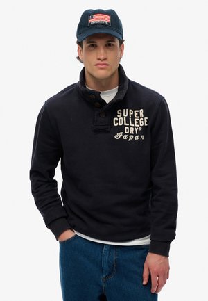 Granatowy sweter z kołnierzykiem i listwą guzikową, z białym napisem "SUPER COLLEGE DRY JAPAN". W połączeniu z niebieskimi dżinsami.