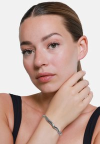 Bracciale in metallo argenteo con un design ondulato, che mostra una superficie liscia e una finitura lucida. Indossato al polso, esalta il tono della pelle.