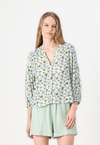 Floral-patterned, lightweight blouse in white with green, blue, and brown flowers, featuring a V-neck and three-quarter sleeves. Paired with mint shorts.