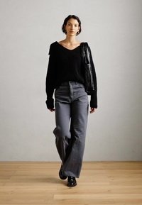 Nő sétál benti térben, fekete pulcsit, szürke, bő szárú nadrágot, fekete cipőt visel, és egy fekete, textúrázott táska lóg a vállán.