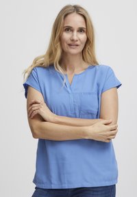Světle modrá blůza s krátkým rukávem, přední kapsou a výstřihem do V. Hladká tkanina s volným střihem, nošena s modrými džíny.
