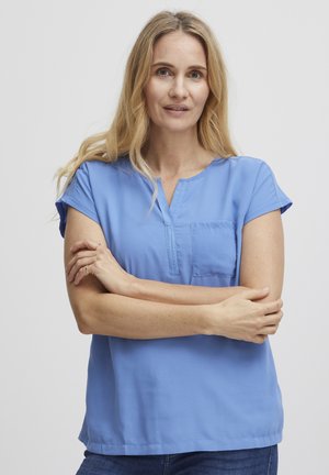 Světle modrá blůza s krátkým rukávem, přední kapsou a výstřihem do V. Hladká tkanina s volným střihem, nošena s modrými džíny.