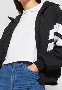 Chaqueta bomber negra con cremallera y capucha, con detalles blancos en las mangas, combinada con una camiseta polo blanca y jeans azules.