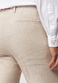 Béžové lněné kalhoty s jemnou texturou a jednou zadní kapsou, doplněné bílou košilí s knoflíky a kulatým límcem.