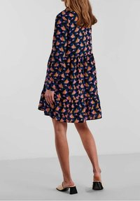 Abito multicolore con un motivo geometrico, maniche lunghe, design a strati e orlo arrotondato. Indossato con scarpe con tacco a blocco open-toe.