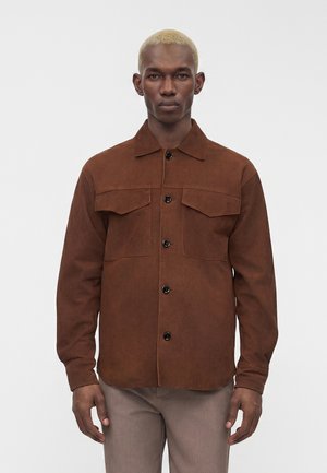 Homme aux cheveux blonds courts portant une veste-chemise marron boutonnée et un pantalon gris clair, debout devant un fond blanc uni.