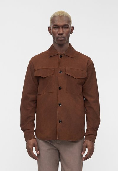 Homme aux cheveux blonds courts portant une veste-chemise marron boutonnée et un pantalon gris clair, debout devant un fond blanc uni.