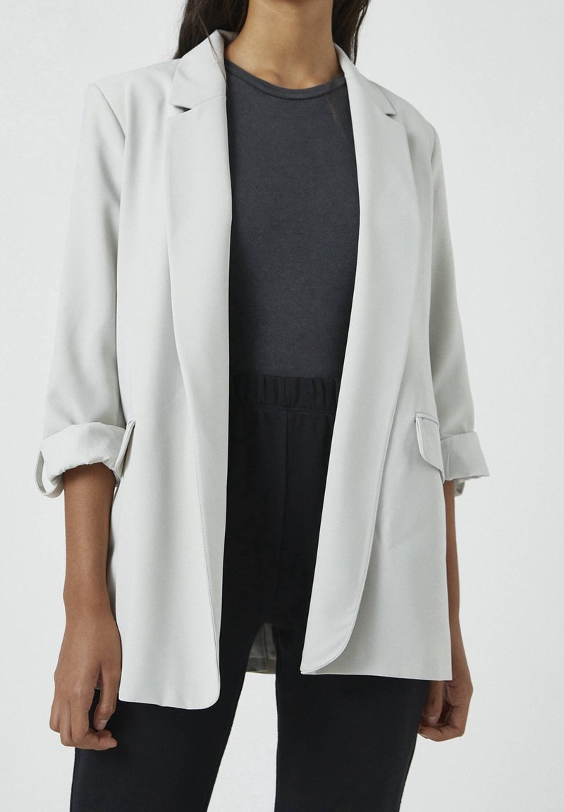 Femme portant un blazer gris clair avec les manches retroussées sur un haut gris foncé et un pantalon noir, vue du cou aux hanches.