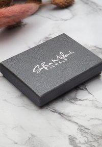 Grå texturerad smyckeslåda med en silverlogotyp på locket, kvadratisk form och släta kanter, placerad på en marmorytan.