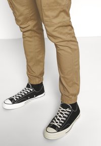 Khaki teplákové kalhoty s elastickými manžetami, spárované s černými plátěnými teniskami s bílou gumovou podrážkou a kontrastními bílými šňůrkami.
