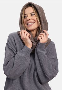 Uśmiechnięta kobieta w szarym swetrze i grubym brązowym kapturze, trzymająca krawędzie kaptura blisko twarzy.