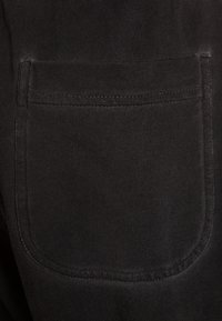 Černá látková kapsa na kalhotách, se čtvercovým tvarem, viditelným prošitím a hladkou strukturou.