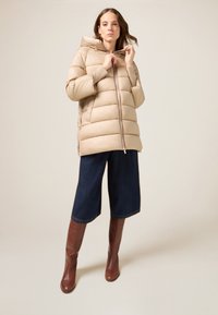 Beżowa kurtka puchowa z kapturem, pikowanym wzorem i zamkiem z przodu. Połączona z ciemnymi denimowymi kulotami i brązowymi kozakiem za kolano.