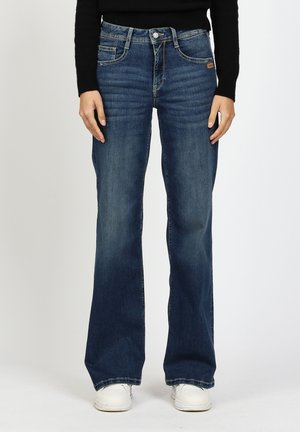 Person trägt dunkelblaue Jeans mit geradem Bein und weiße Sneakers, steht mit entspannten Händen an den Seiten vor einem schlichten Hintergrund.