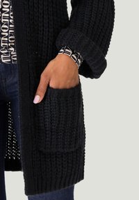 Schwarze Strickjacke mit grober Textur, ausgestattet mit einer großen Vordertasche und aufgeschlagenen Ärmelbündchen, über einem gemusterten Oberteil getragen.