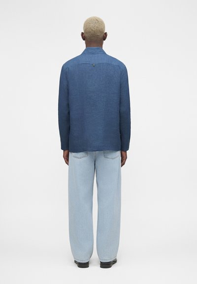 Άτομο με κοντά ξανθά μαλλιά, που φοράει μπλε μπλούζα με μακριά μανίκια και ανοιχτό μπλε τζιν με φαρδιά πόδια, στέκεται γυρισμένο με την πλάτη σε μονόχρωμο φόντο.