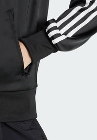 Crna sportska jakna s bijelim prugama na rukavima, elastičnim manžetama, bočnim džepovima sa zatvaračem i glatkom, sjajnom teksturom.