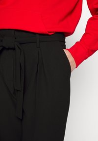 Černé kalhoty s páskem, se sklady a bočními kapsami, v kombinaci s červenou mikinou s klokankou a žebrovanými manžetami.