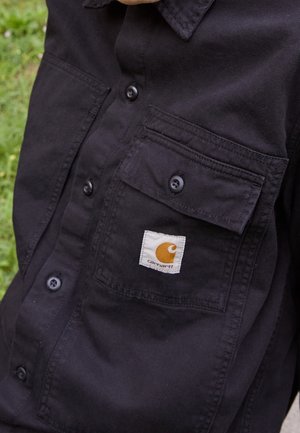 Close-up of black button-up shirt with two chest pockets, one featuring a white and yellow Carhartt logo patch.