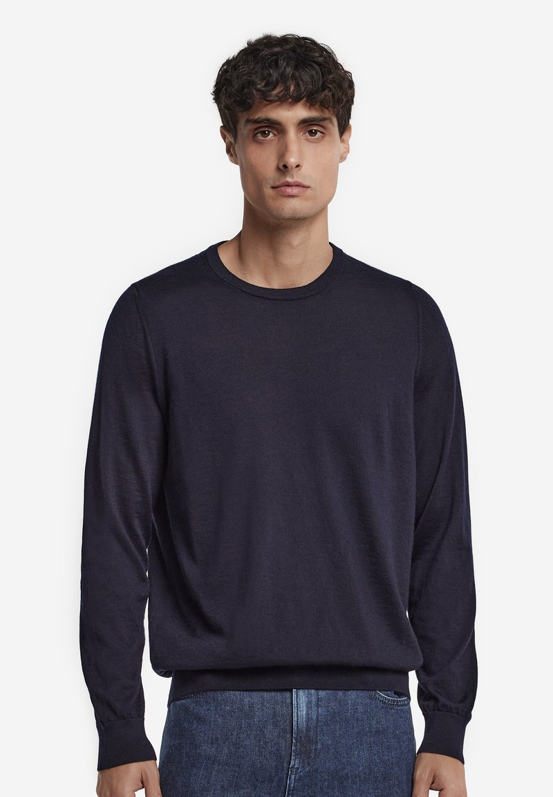Ung mand med kort krøllet mørkt hår iført en ensfarvet mørkeblå langærmet sweater og blå jeans, stående mod en hvid baggrund.