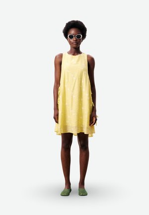 Femme aux cheveux courts et bouclés porte des lunettes de soleil blanches, une robe jaune sans manches et des chaussures vertes sans lacets, debout devant un fond uni.