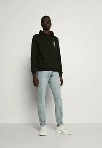 Hoodie preto com uma estampa na frente, combinado com jeans de denim azul claro e ténis brancos. O hoodie apresenta um cordão e punhos canelados.