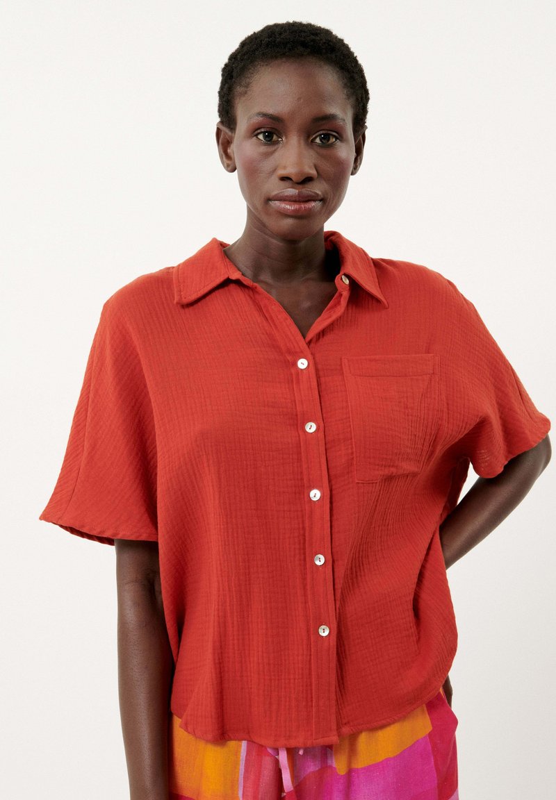 Une chemise à manches courtes, coupe décontractée, en tissu rouge texturé, avec un devant à boutons, un col et une poche sur le côté gauche.